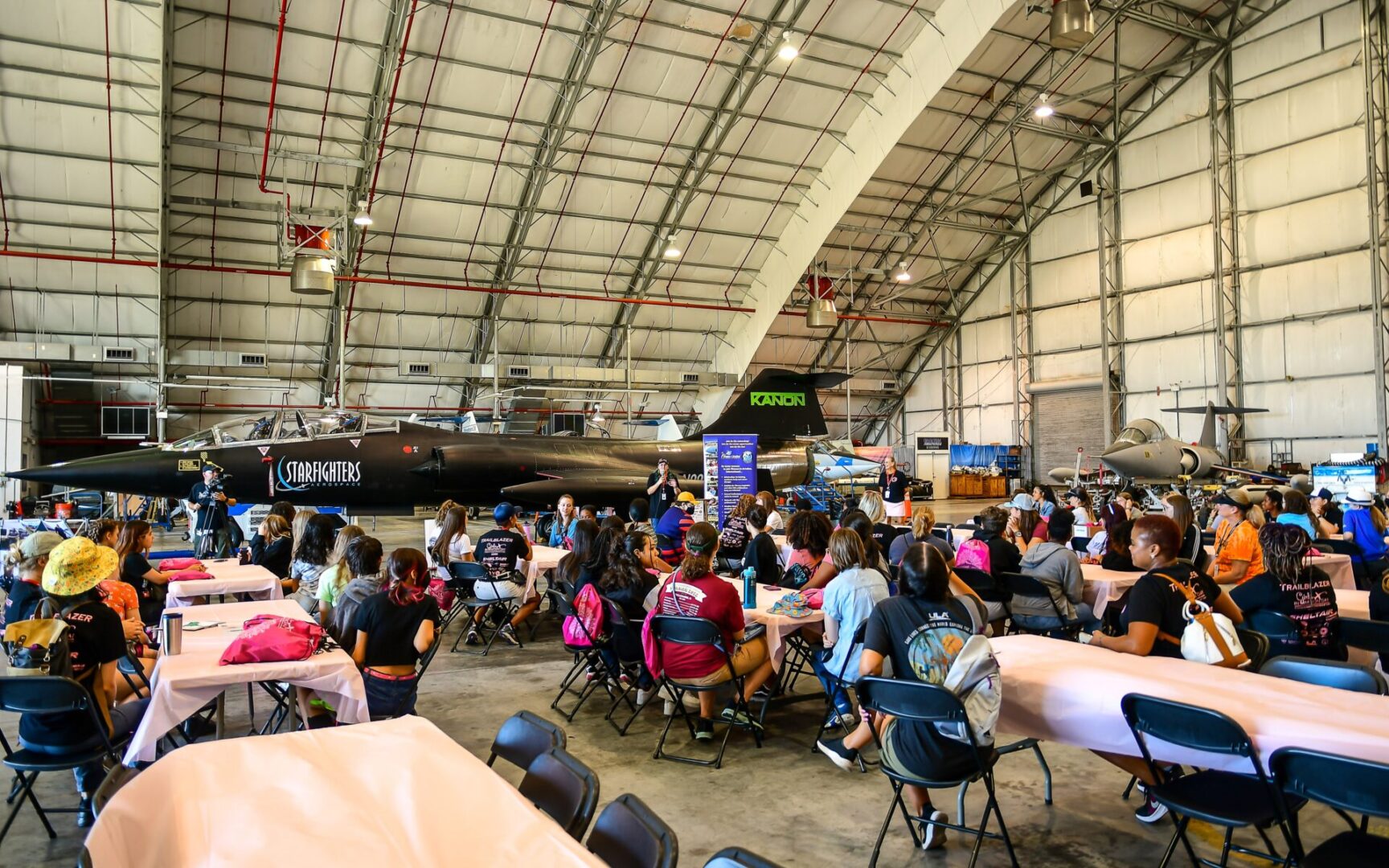 A group of people sitting around tables in an airplane hangar.