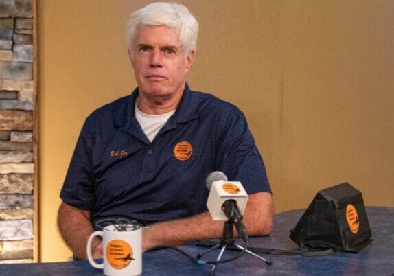 Man seated at a table with microphones.